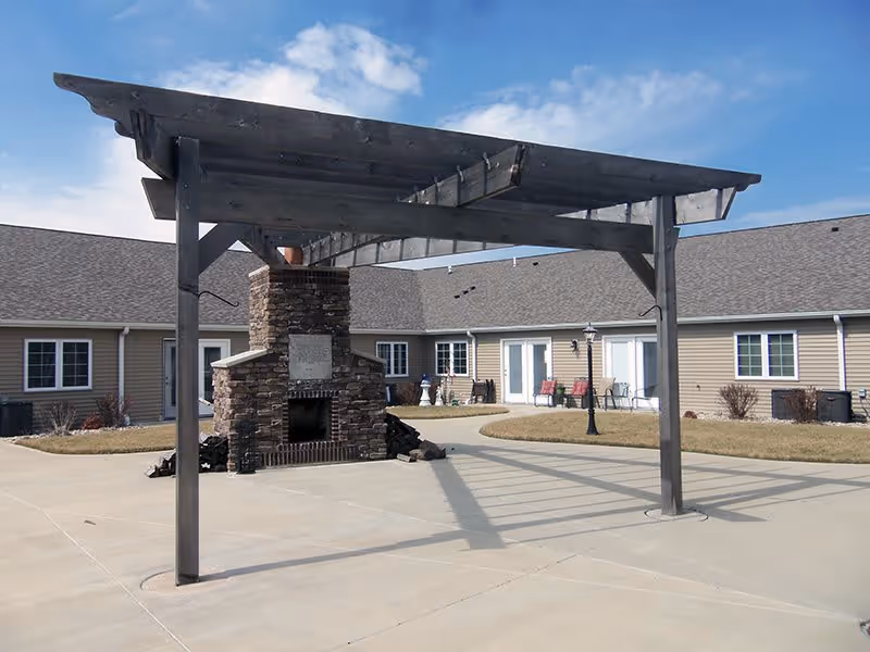 Outdoor courtyard area with a wooden pergola and a stone fireplace. Surrounding the courtyard are single-story beige buildings with multiple windows and doors. The sky is partly cloudy.