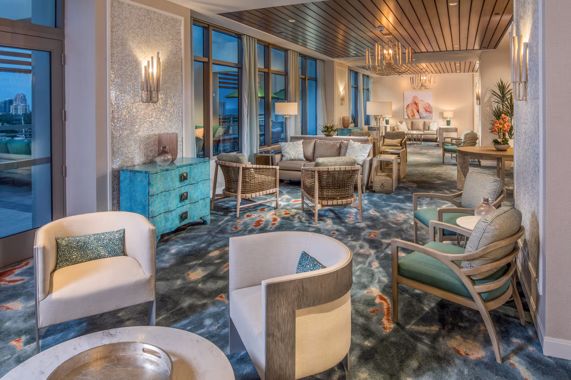 A spacious and elegantly decorated senior living common area with multiple seating arrangements including armchairs, sofas, and side tables. The room features large windows with curtains, modern wall sconces, chandeliers, and a patterned carpet. There are decorative plants and artwork on the walls, creating a comfortable and inviting atmosphere.