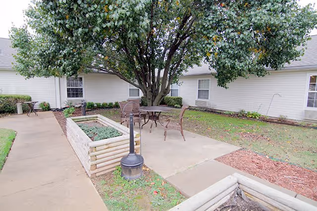 Outdoor courtyard area with a large tree in the center, surrounded by a concrete patio with metal chairs and tables. Raised garden beds made of wooden logs are visible along the walkway. The courtyard is enclosed by a single-story building with white siding and several windows.