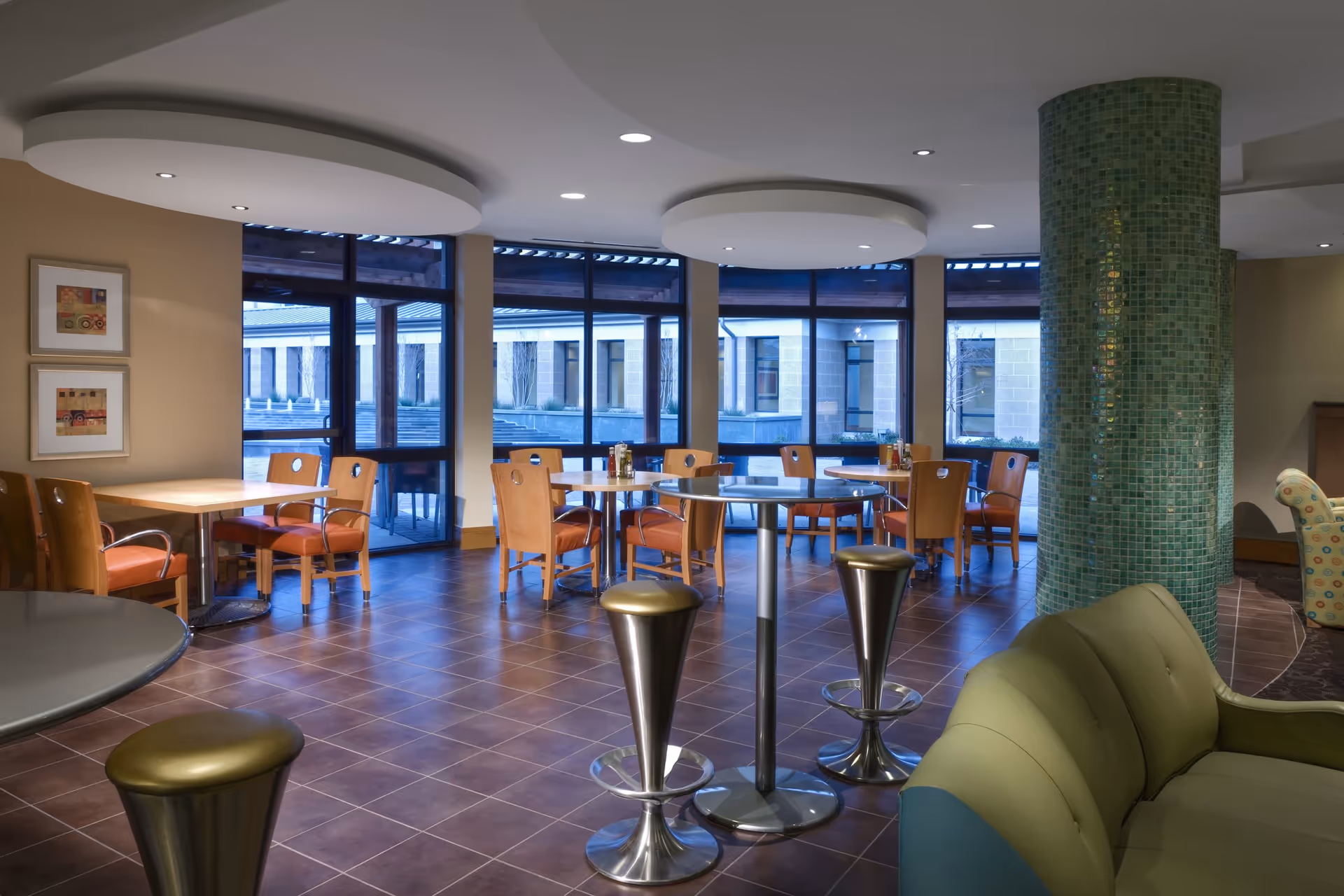 A modern dining area with several wooden tables and chairs arranged near large windows that look out onto an outdoor courtyard. The room features a green mosaic tiled column, circular ceiling light fixtures, and a green cushioned sofa in the foreground. The floor is tiled with brown tiles.