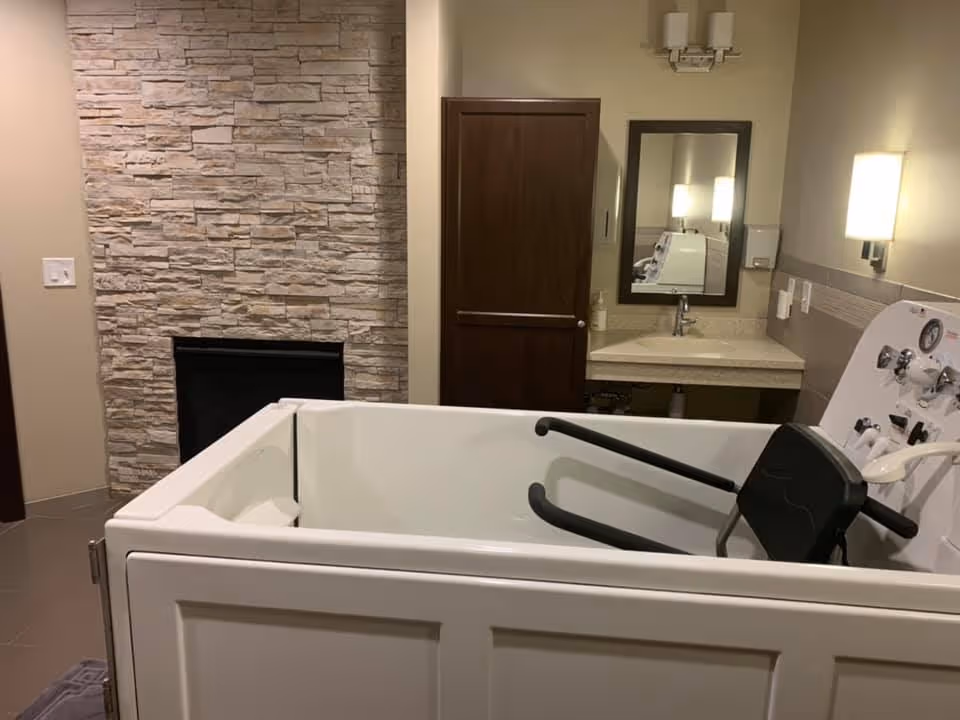 A bathroom designed for assisted living with a walk-in bathtub featuring safety handles and a seat. The room has a stone accent wall with a fireplace, a wooden door, a sink with a mirror above it, and wall-mounted lights providing warm illumination.