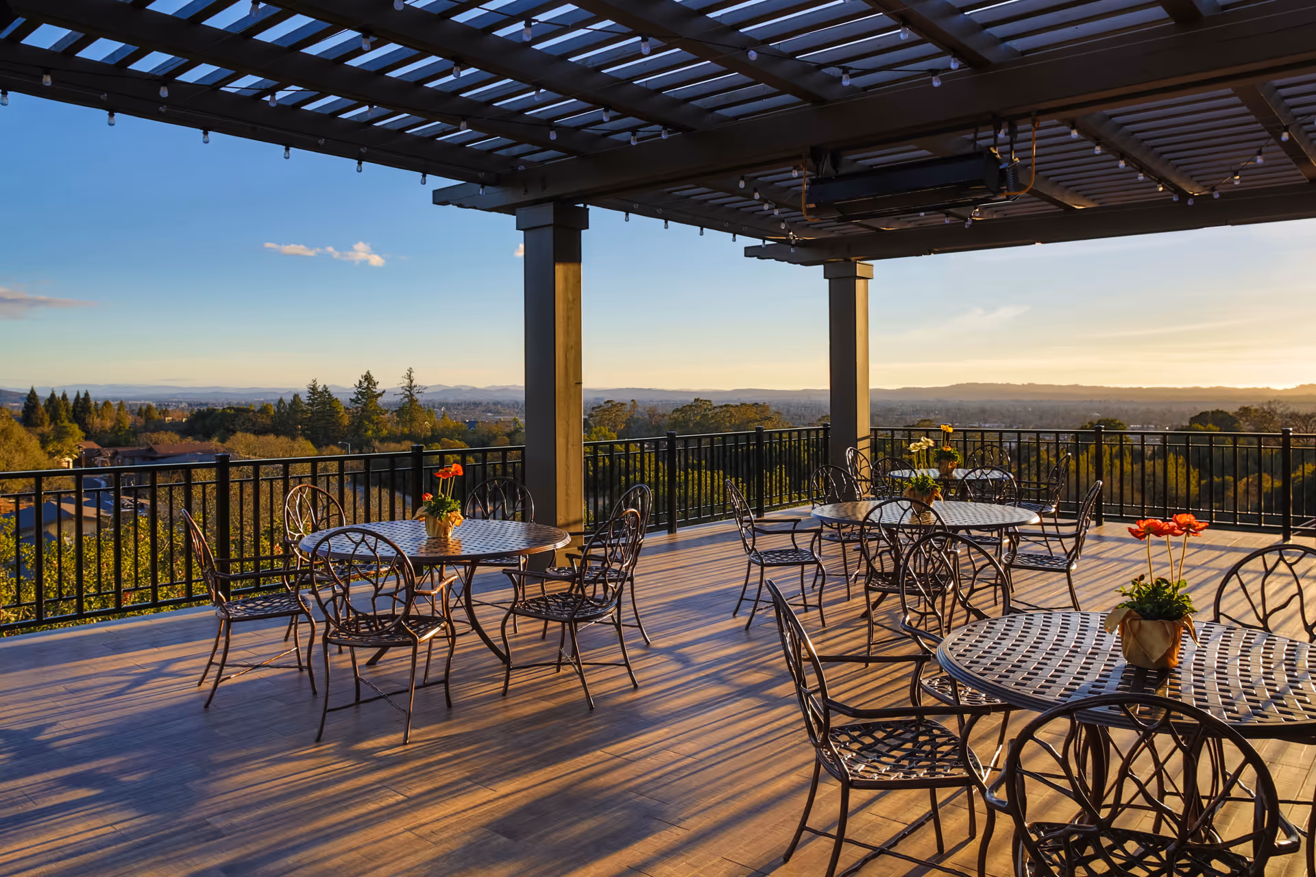 Outdoor patio area with metal tables and chairs under a pergola roof, decorated with small potted plants with flowers, overlooking a scenic view of trees and distant hills during sunset.