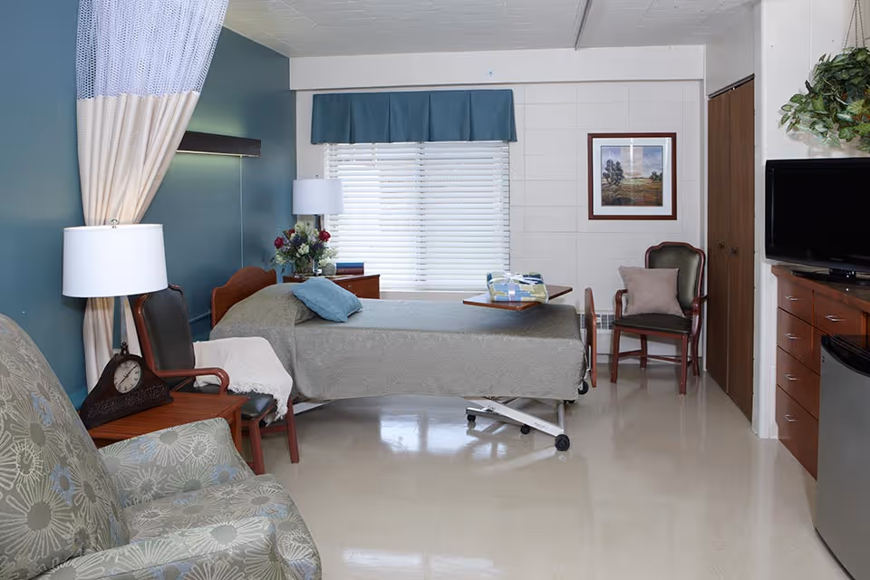 A senior living facility bedroom with a hospital-style bed covered in a gray blanket and a blue pillow. The room has a large window with blinds and a blue valance, a wooden nightstand with a lamp and flowers, two chairs with cushions, a small table on wheels over the bed, a television on a wooden dresser, and a small refrigerator. The walls are white with a framed picture and a hanging plant.