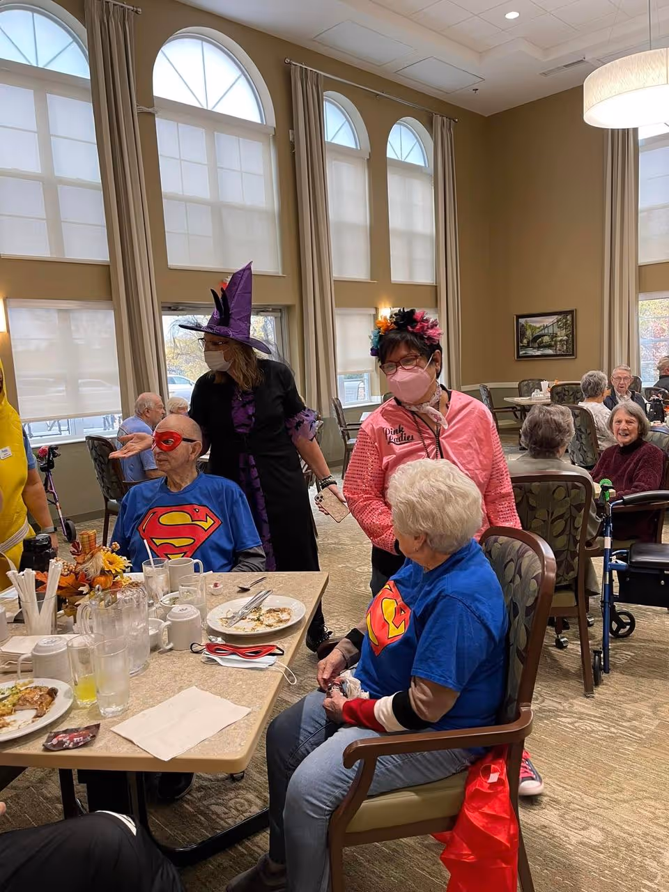 A group of elderly people and staff members in a dining room at Prairie Pointe Assisted Living and Memory Care. Two elderly individuals are seated at a table wearing blue shirts with a Superman logo, one also wearing a red mask. Two staff members are standing nearby, one dressed in a witch costume with a purple hat and the other wearing a pink jacket and a colorful flower headband. Other residents are seated at tables in the background, engaged in conversation. The room has large arched windows with beige curtains and warm lighting.