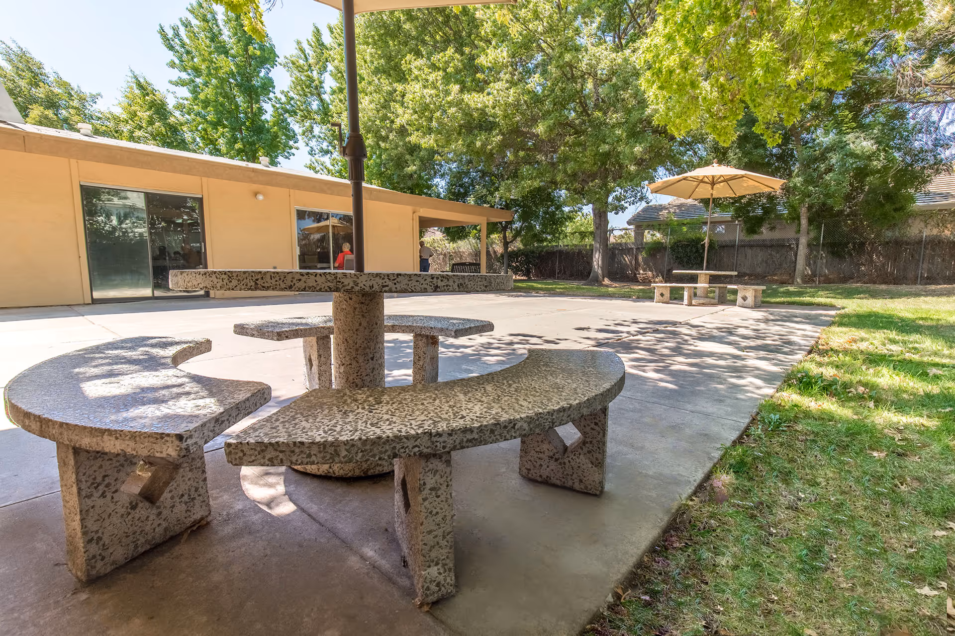 Outdoor patio area with circular stone picnic tables and benches under a sunny sky. There are trees providing shade and a building with large windows in the background.