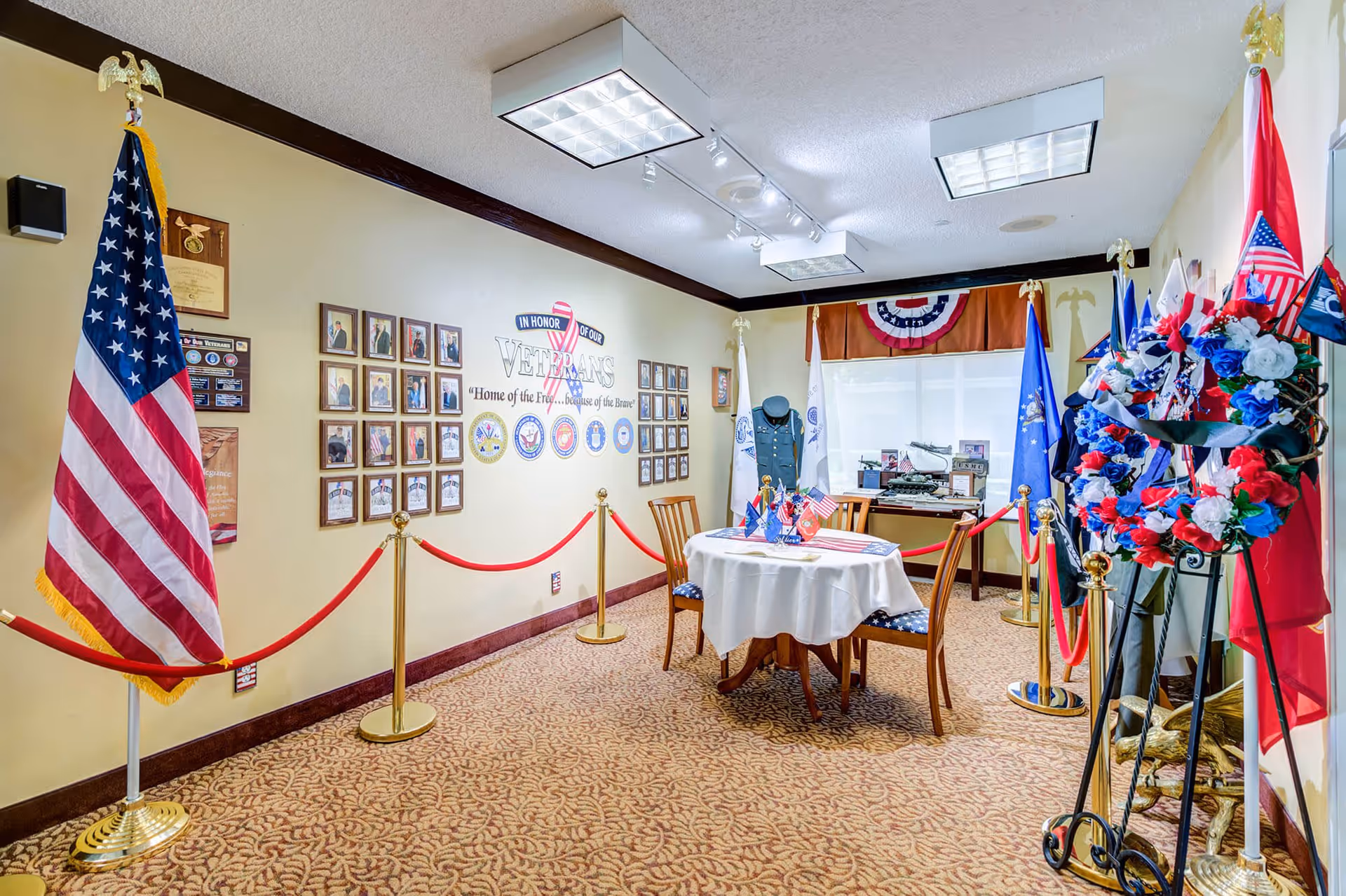 A room dedicated to honoring veterans with American flags, military insignia, framed photos and plaques on the walls, a table with small flags, and a wreath decorated with red, white, and blue flowers. The room is carpeted and has bright ceiling lights.