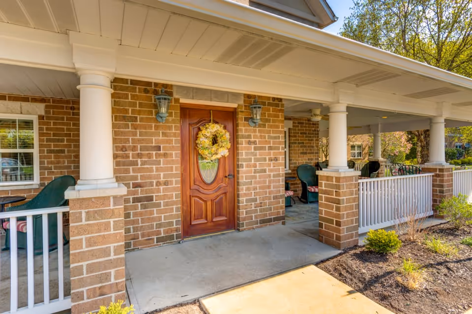 Covered brick front porch with a wooden entry door adorned with a yellow floral wreath, white columns, and outdoor seating.