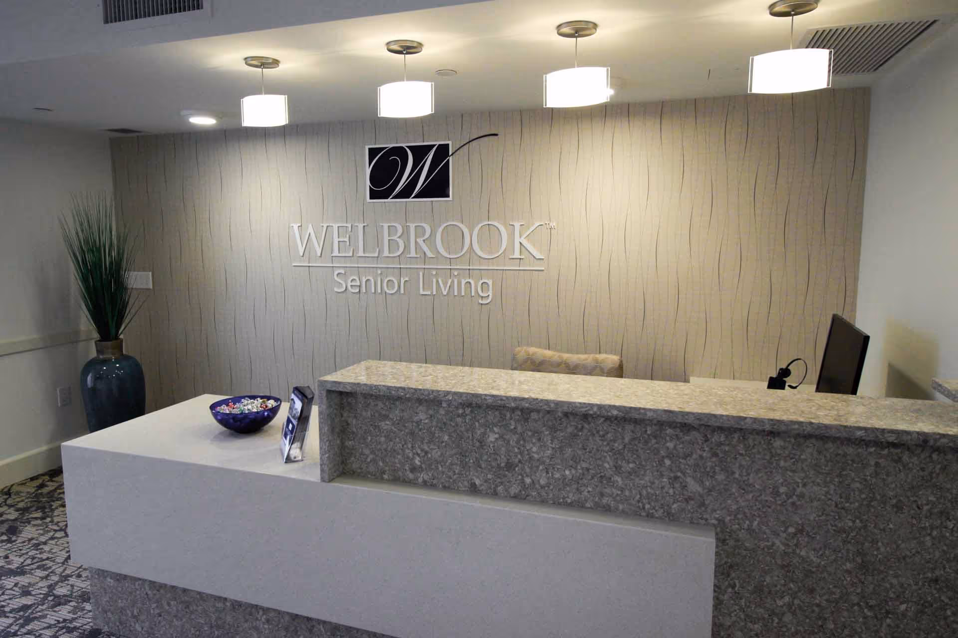 Reception desk area at Welbrook Senior Living facility with a granite countertop, a bowl of candy, informational brochures, a computer monitor, and a tall vase with green plants against a textured wall with the Welbrook Senior Living logo.