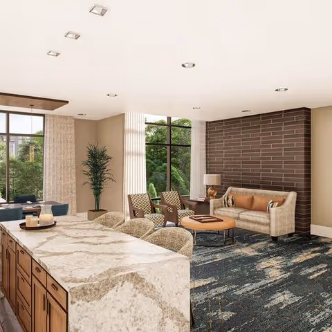 A spacious and modern living room area with a large marble countertop island in the foreground featuring four woven chairs. The room includes a beige sofa with cushions, two patterned armchairs, a round coffee table, a floor lamp, and a tall green plant near large windows with curtains that let in natural light. The walls are light-colored with a dark brick accent wall behind the sofa, and the floor is covered with a patterned carpet.