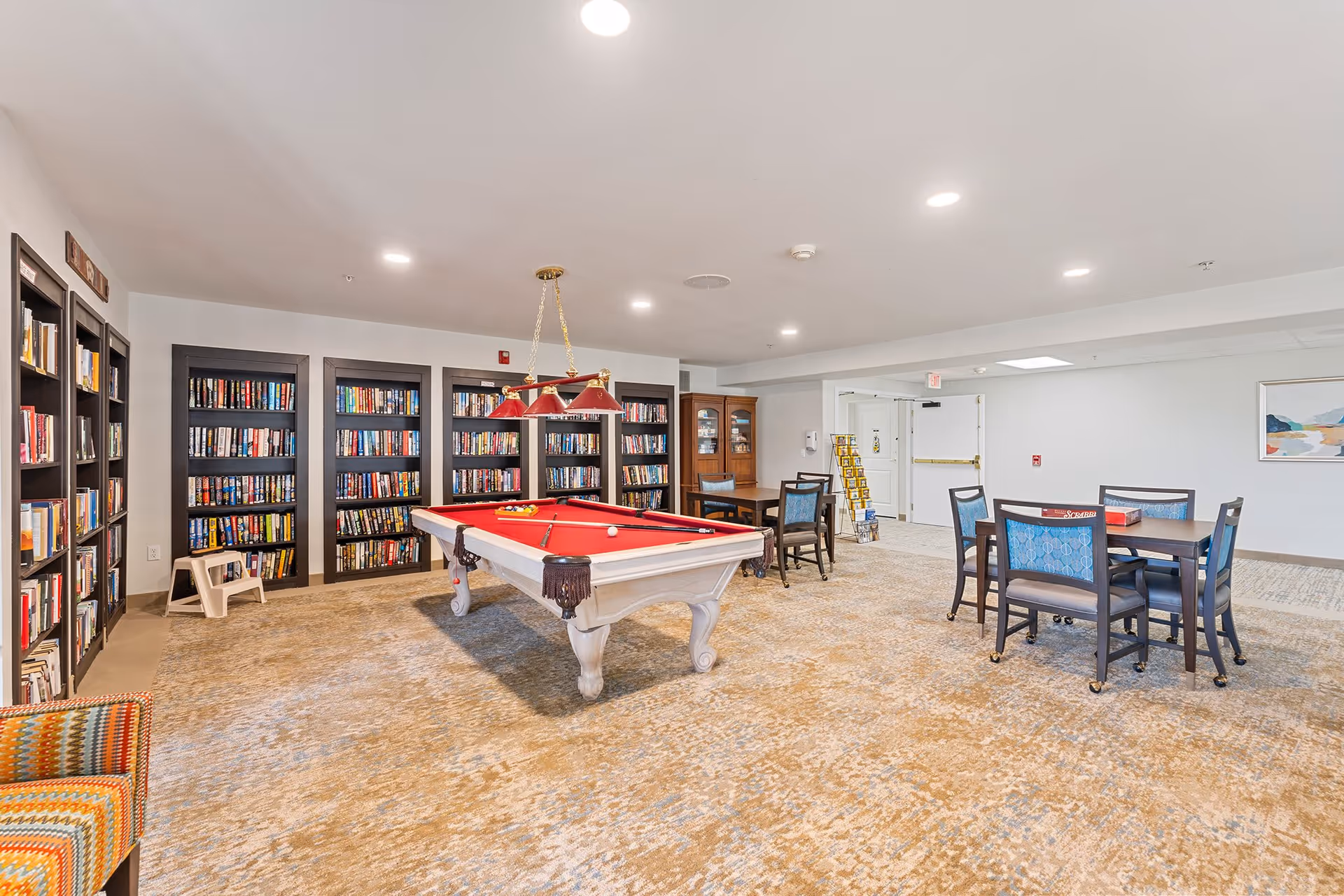 A spacious recreational room with a red felt pool table in the center, surrounded by bookshelves filled with books along the left wall. There are two tables with chairs on the right side, one with a board game on it. The room has beige carpeting, white walls, and recessed ceiling lights.