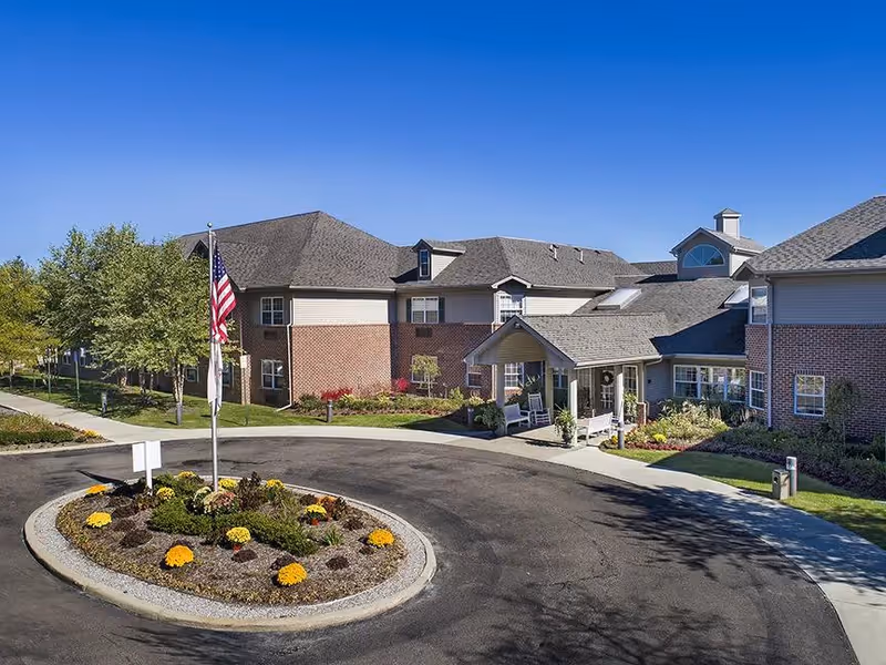 Exterior view of a senior living facility building with a circular driveway and a landscaped roundabout featuring yellow flowers and an American flag. The building has a combination of brick and siding with multiple windows and a covered entrance. Trees and a clear blue sky are visible in the background.