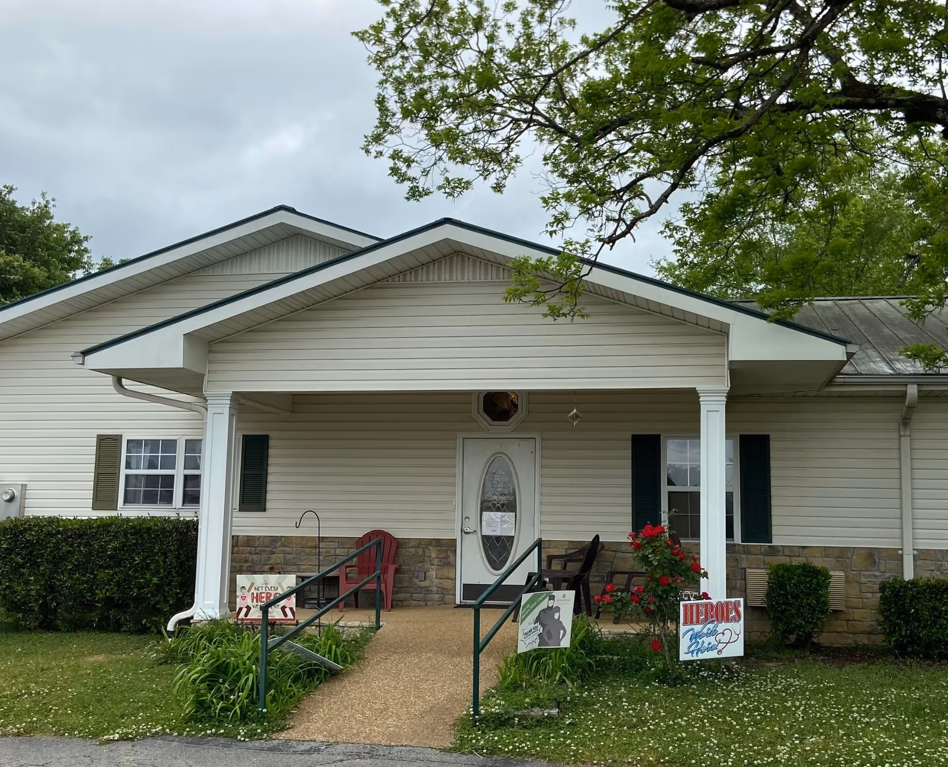 Front exterior view of a single-story building with beige siding and a covered porch supported by white columns. There are two chairs on the porch, a pathway with handrails leading to the front door, and several signs in the yard including one that says 'Heroes Work Here'. Green bushes and a tree with green leaves surround the building under a cloudy sky.