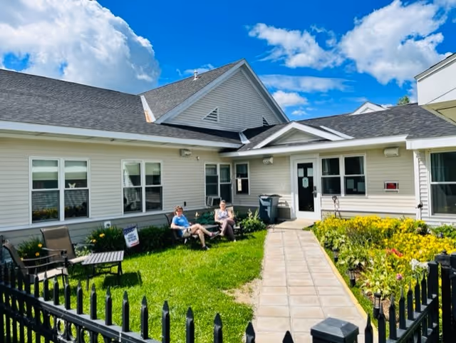 Outdoor courtyard area of a senior living facility with two people sitting on chairs on a grassy lawn. The building is single-story with beige siding and multiple windows. There is a paved walkway leading to a door, surrounded by green plants and flowers. The sky is blue with some clouds.