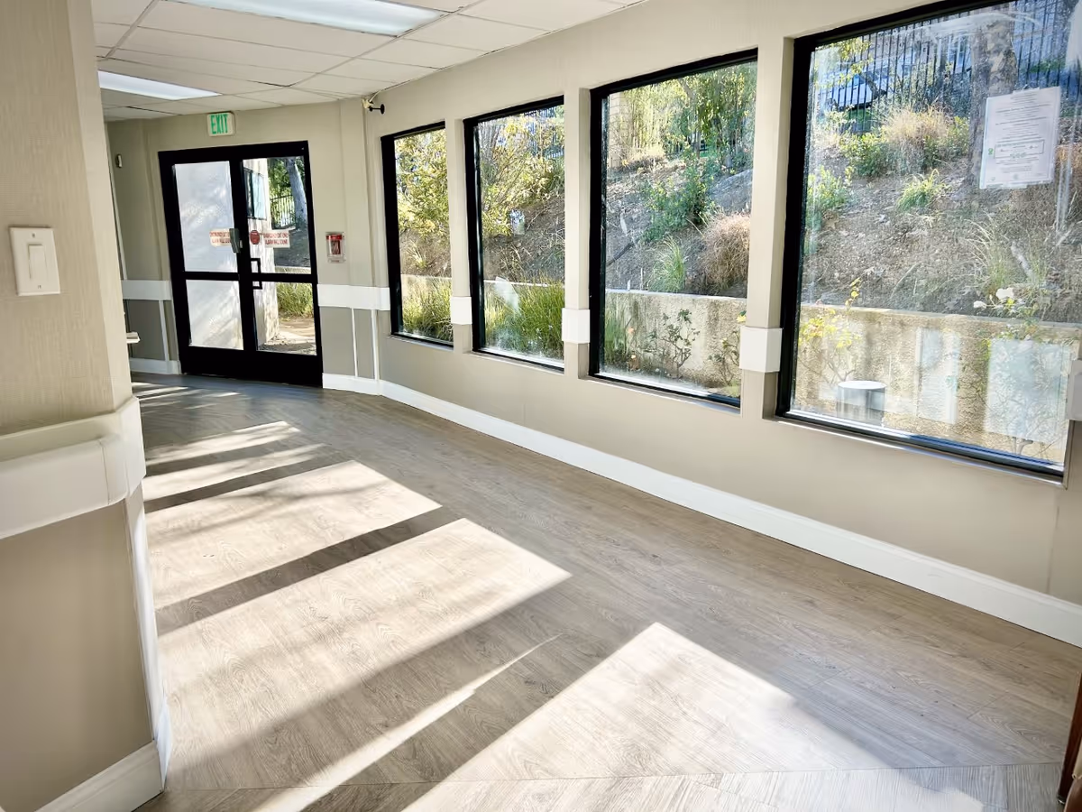 Sunlit interior corridor with large windows along the right wall and an exit door at the far end.