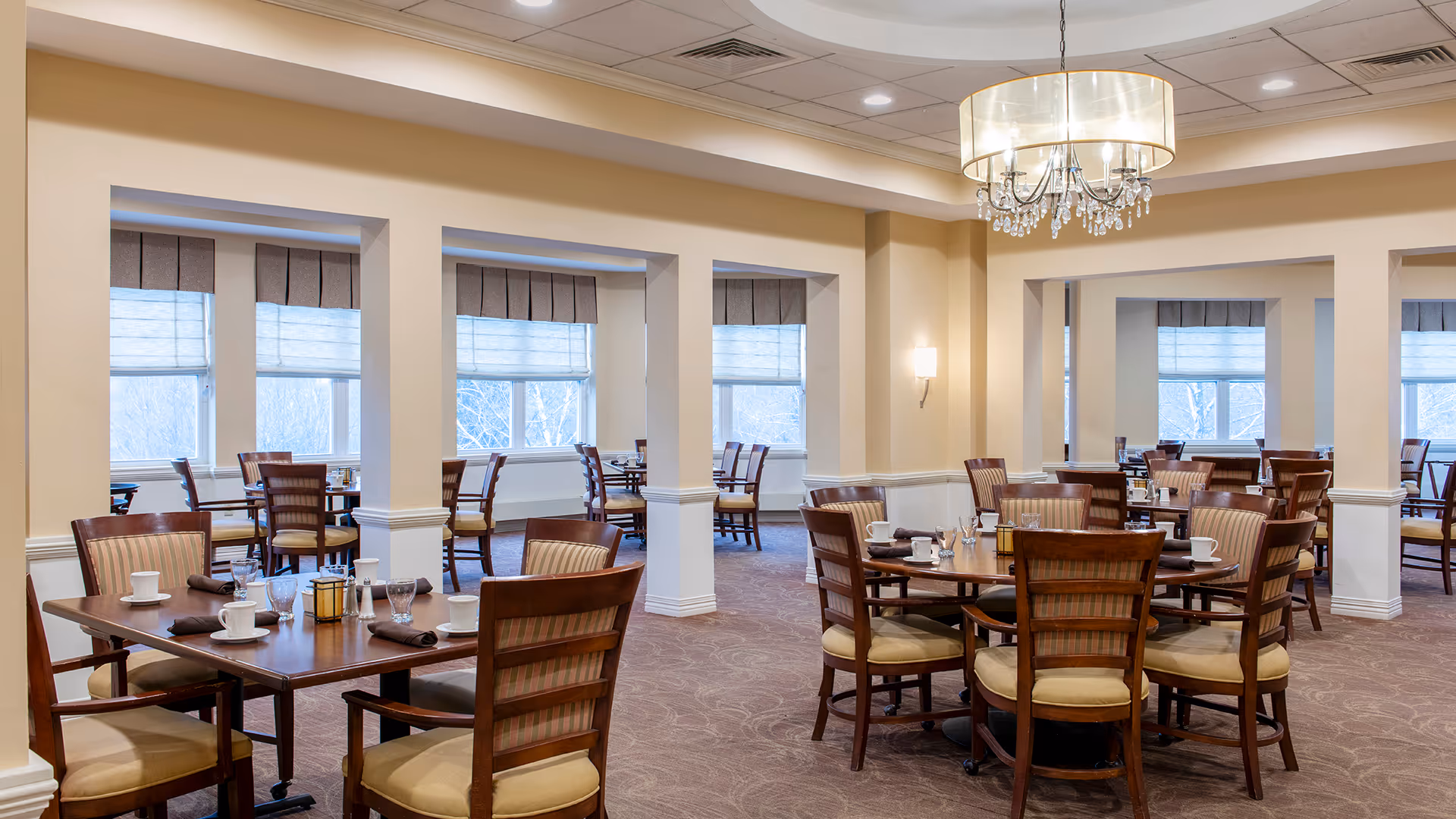 A spacious dining room with multiple wooden tables and chairs arranged neatly. Each table is set with white cups, glasses, and napkins. The room has large windows with blinds allowing natural light to enter. A chandelier hangs from the ceiling, and the walls are painted in a light beige color.