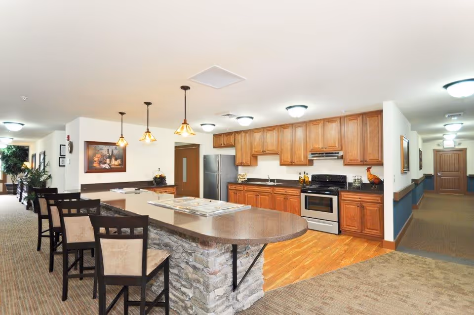 Interior view of a senior living facility kitchen and dining area with wooden cabinets, stainless steel refrigerator and stove, a stone-faced island with a brown countertop, and four high chairs. The area is well-lit with ceiling lights and pendant lights above the island. A hallway extends from the kitchen area with framed artwork on the walls.