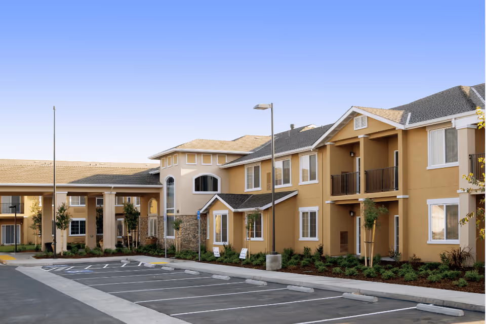 Exterior view of Manteca Manor, a two-story senior living facility with beige walls, white trim, and a stone accent near the entrance. The building has multiple windows and balconies, surrounded by landscaped greenery and an empty parking lot in front under a clear blue sky.