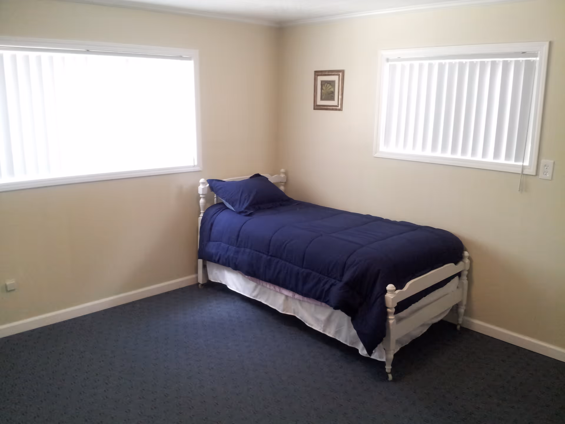 A simple bedroom with a single white wooden bed frame, a navy blue comforter and pillow, beige walls, blue carpet, two windows with white vertical blinds, and a small framed picture on the wall.
