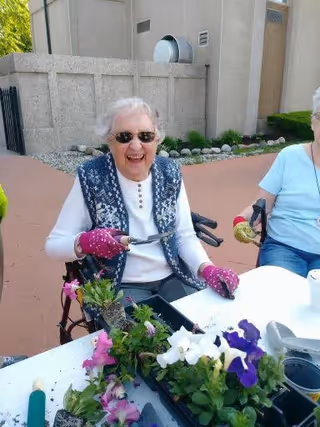 An elderly woman wearing sunglasses and pink gardening gloves is smiling while holding a small gardening tool. She is seated outdoors at a table with various flowers and gardening supplies. Another person is partially visible sitting next to her.