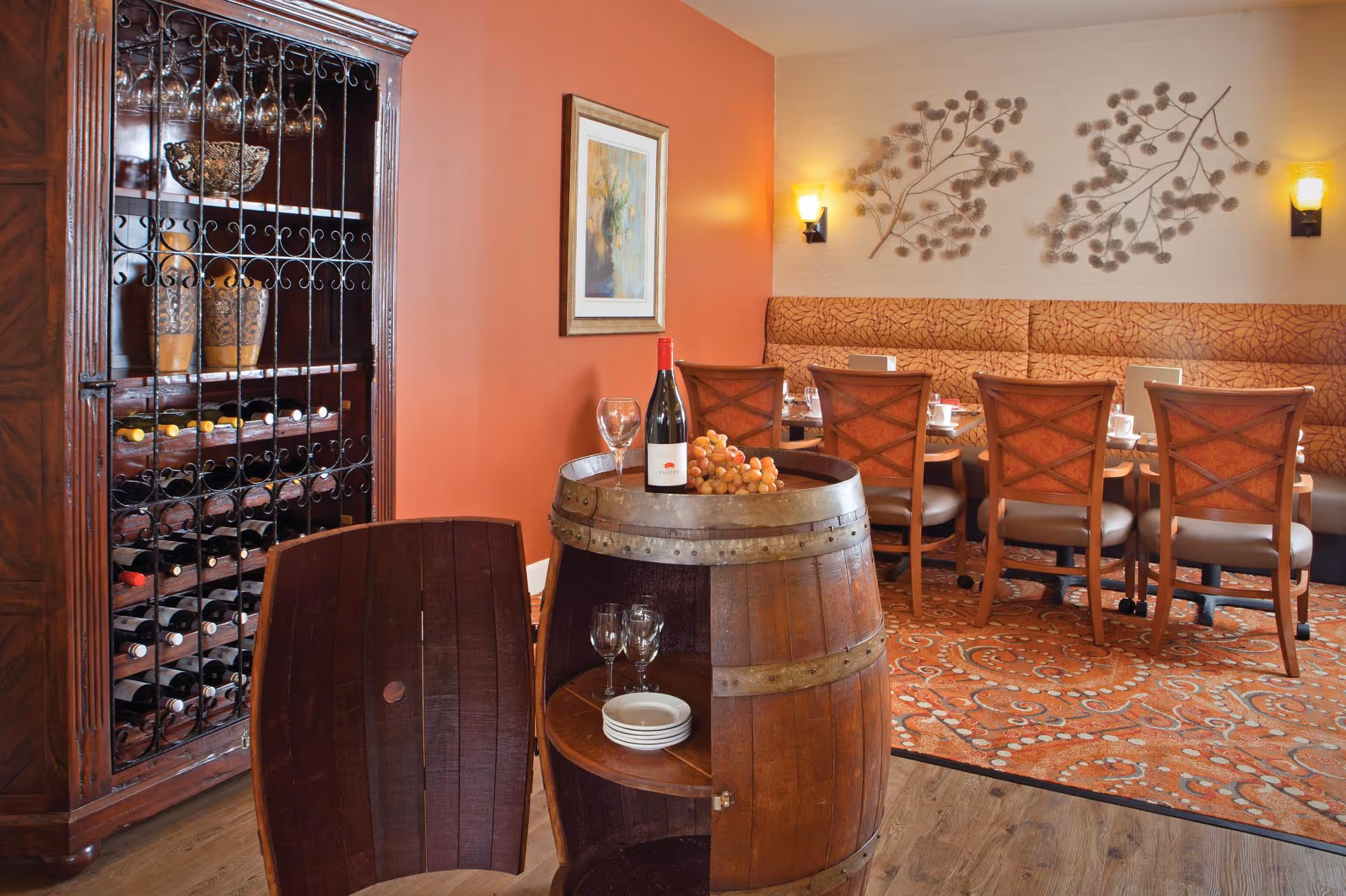 Dining room with a wine cabinet and a wooden barrel table topped with a bottle of wine and glasses, and a banquette with dining chairs.