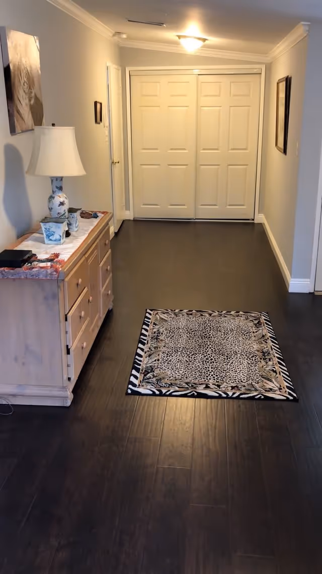 A long interior hallway with dark wood floors, a small patterned rug, a wooden dresser with a lamp on the left, and closed double doors at the far end.