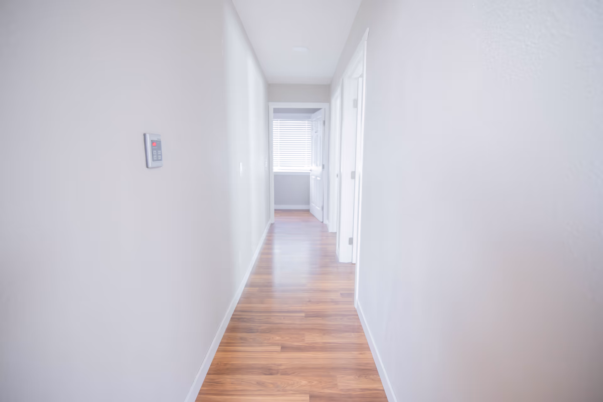 A bright, narrow hallway with light-colored walls and wooden flooring, featuring a thermostat on the left wall and several white doors on the right side, leading to other rooms. Natural light is coming from a window at the end of the hallway.