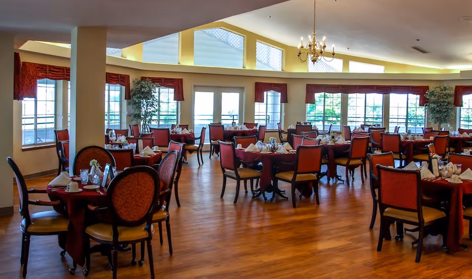 Spacious dining room with multiple round tables covered in red tablecloths, each set with white napkins, glasses, and cups. The room has large windows with red valances, wooden flooring, and a chandelier hanging from the ceiling.
