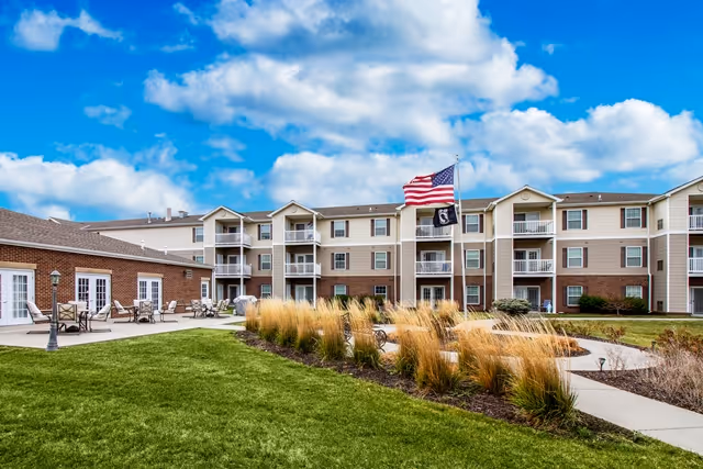 Exterior view of a multi-story retirement community building with balconies, a well-maintained lawn, ornamental grasses, a paved walkway, and an outdoor seating area with tables and chairs. Two flags, including an American flag, are flying on flagpoles in front of the building under a partly cloudy blue sky.