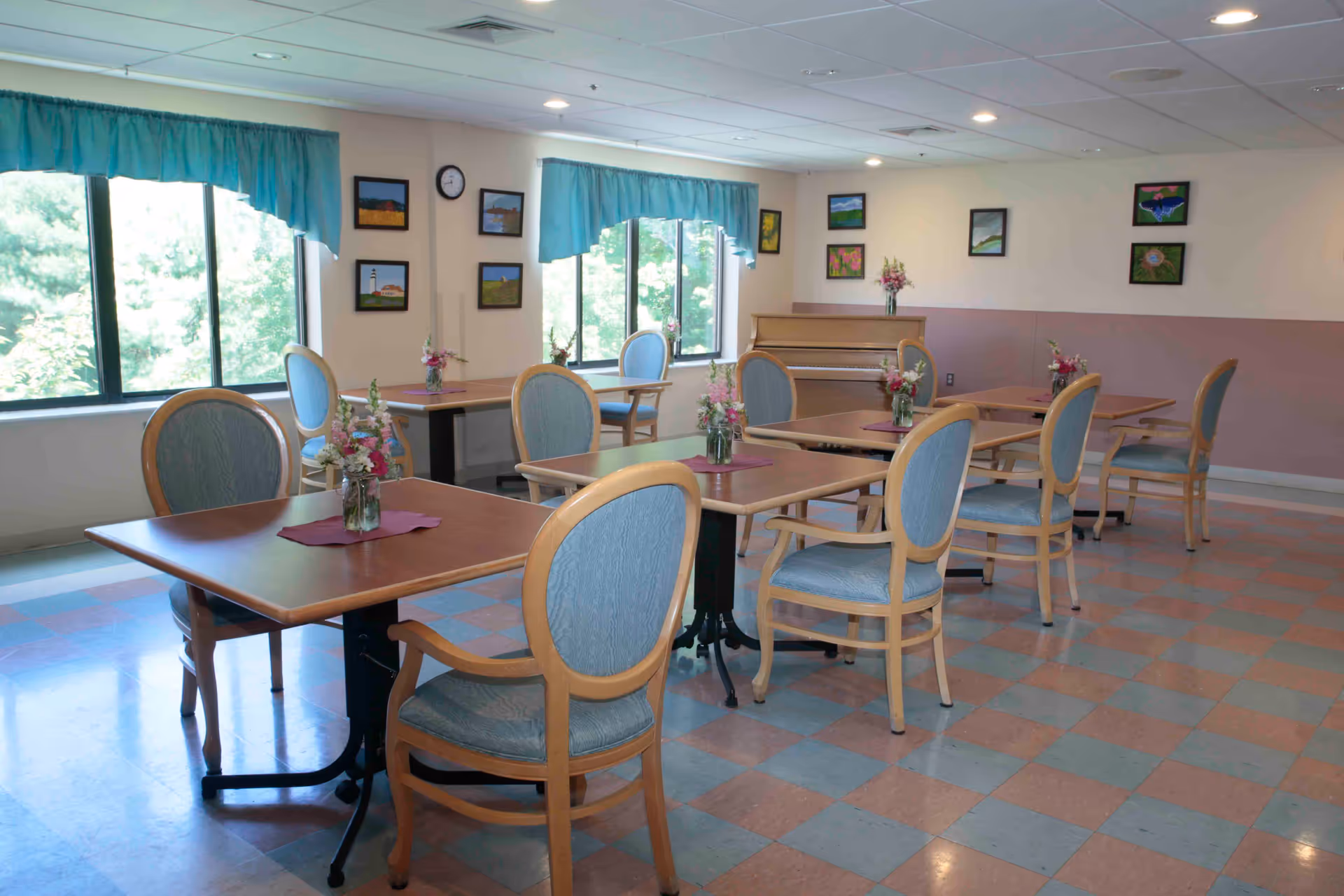 Bright dining room with wooden tables and cushioned chairs arranged with small floral centerpieces, windows with teal valances, and framed pictures on the walls.