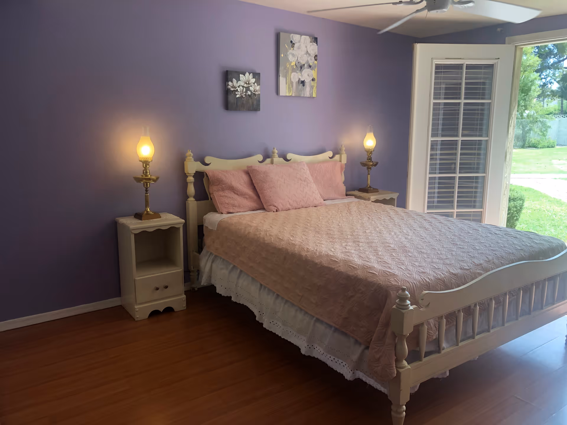 A bedroom with a cream-colored wooden bed frame and a pink quilted bedspread with matching pillows. There are two bedside tables, each with a vintage-style lamp emitting a warm glow. The walls are painted purple and decorated with two floral paintings. A ceiling fan is visible above, and a glass door with white blinds opens to a green outdoor area.
