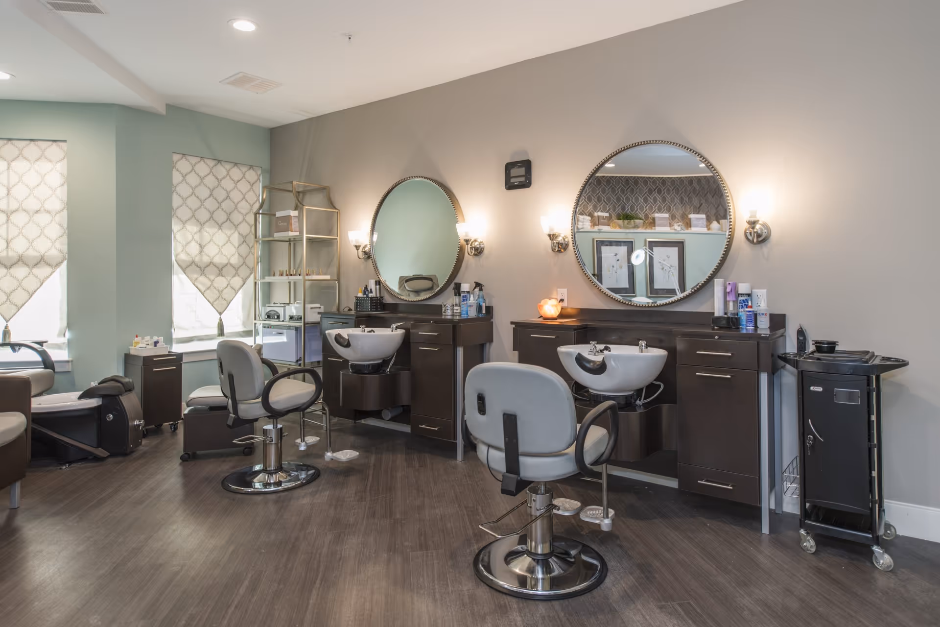 Interior of a salon area with two styling stations, each featuring a round mirror, a wash basin, and a salon chair. The room has light-colored walls, two windows with patterned blinds, and a shelving unit with various salon products. There is also a rolling cart with supplies on the right side.
