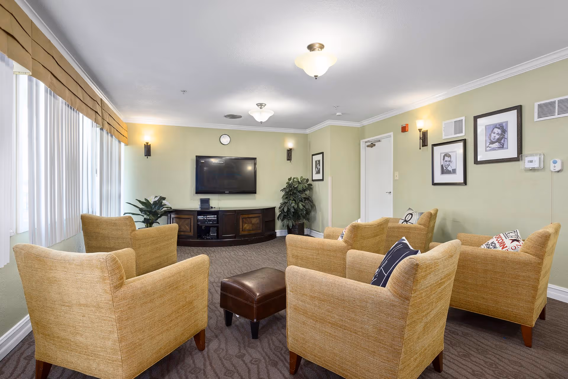 A cozy living room area with six beige armchairs arranged in a semi-circle around a small brown ottoman. A flat-screen TV is mounted on a light green wall above a dark wooden cabinet. The room has carpeted flooring, two potted plants, framed black and white photos on the walls, and large windows with vertical blinds and a valance.