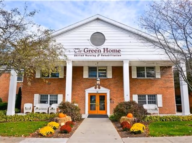 Front exterior view of The Green Home, a senior nursing and rehabilitation facility. The building has a brick facade with white columns and trim, orange double doors at the entrance, and landscaping with colorful flowers and pumpkins along the walkway. Trees flank the building on both sides.
