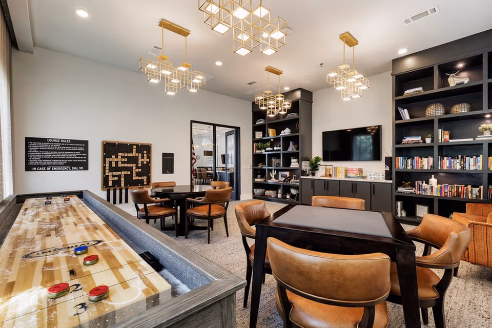 Bright community lounge with a shuffleboard, card/game tables, leather chairs, bookshelves, and a wall-mounted TV.