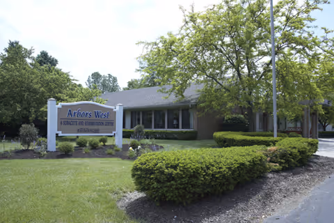 Exterior view of Arbors West, a graduate and rehabilitation center, showing a single-story building surrounded by green bushes, trees, and a well-maintained lawn under a partly cloudy sky.