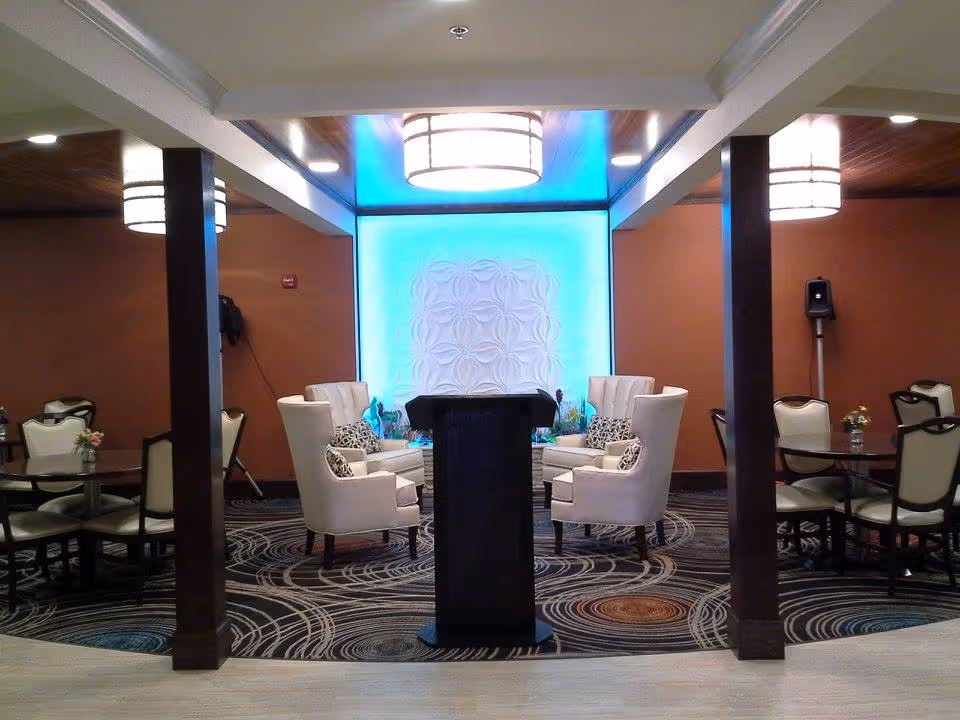 A cozy interior common area with a central podium, surrounded by four white armchairs with patterned cushions. The room features round tables with chairs on either side, decorative lighting fixtures on the ceiling, and a blue illuminated wall panel with a floral pattern in the background.
