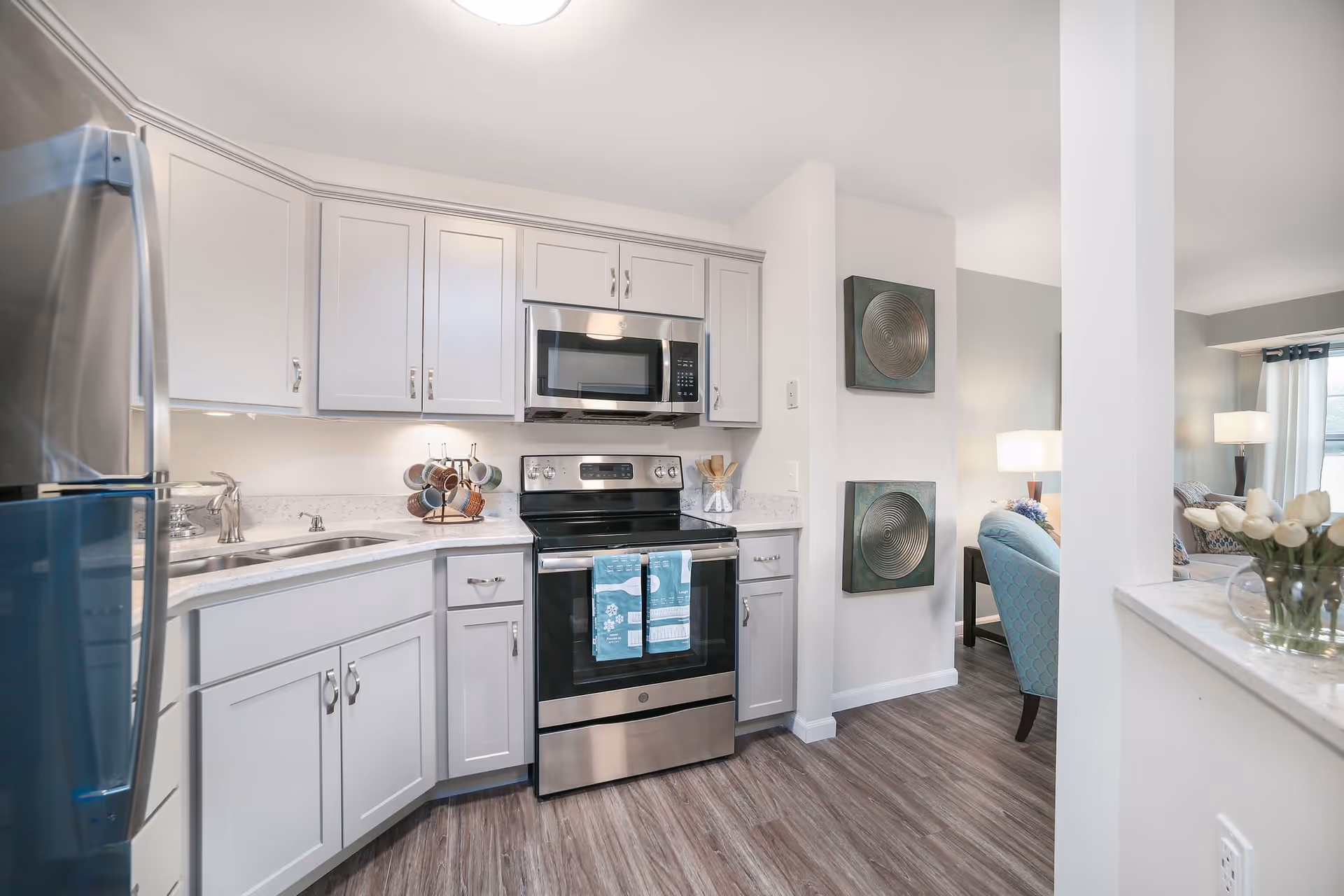 Modern kitchen with white cabinets, stainless steel refrigerator, stove, and microwave. The kitchen has a marble countertop with a sink and a coffee mug holder. Adjacent to the kitchen is a living area with a blue armchair, lamps, and wall art.