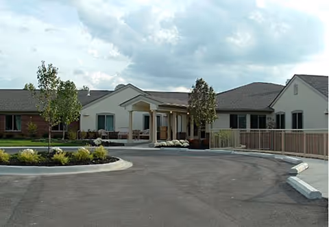 Single-story senior living facility entrance with a covered porte-cochere, circular driveway and landscaped beds under a cloudy sky.