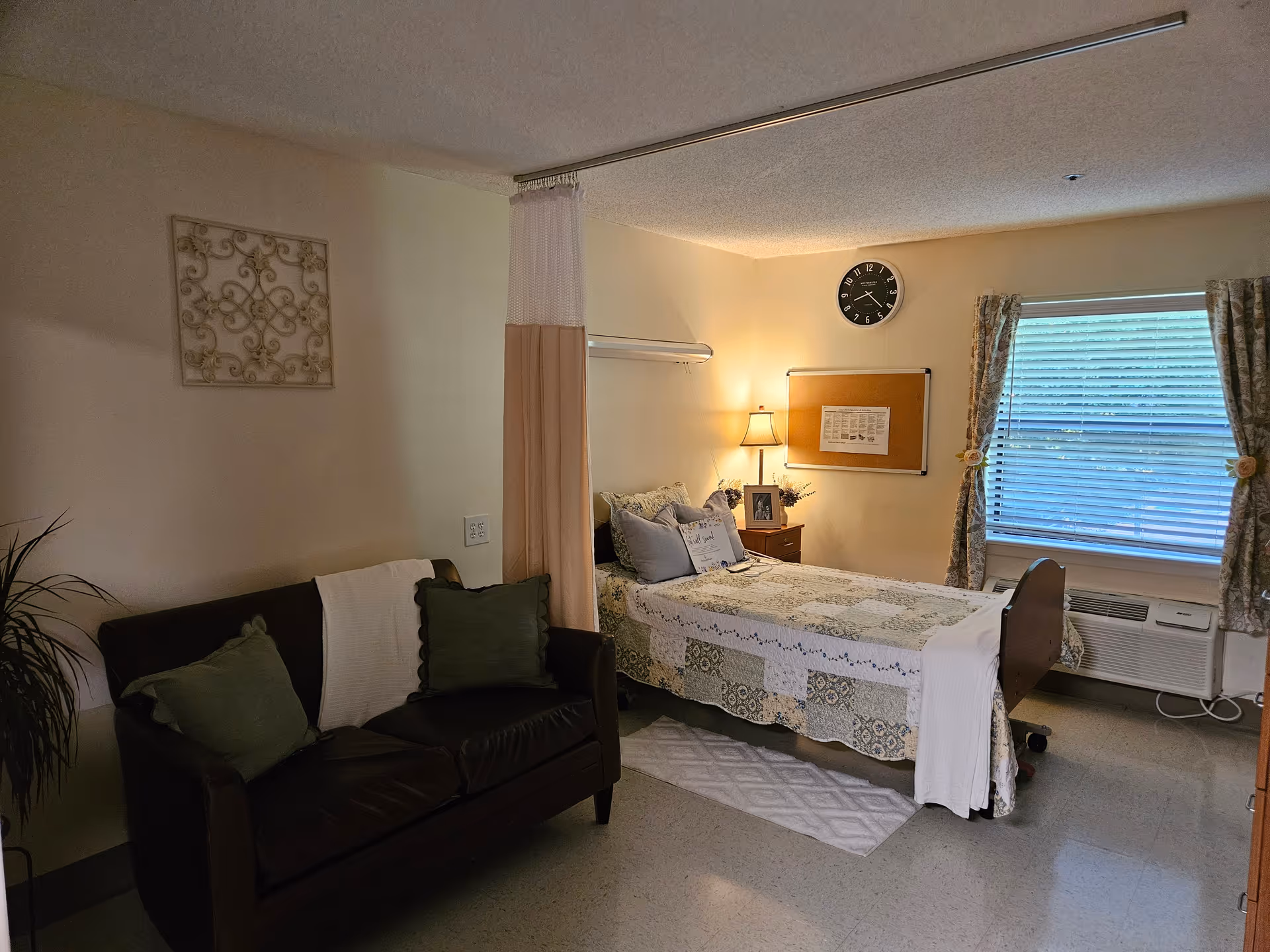 Bedroom in a senior living facility with bed, sofa, nightstand, lamp, and window.