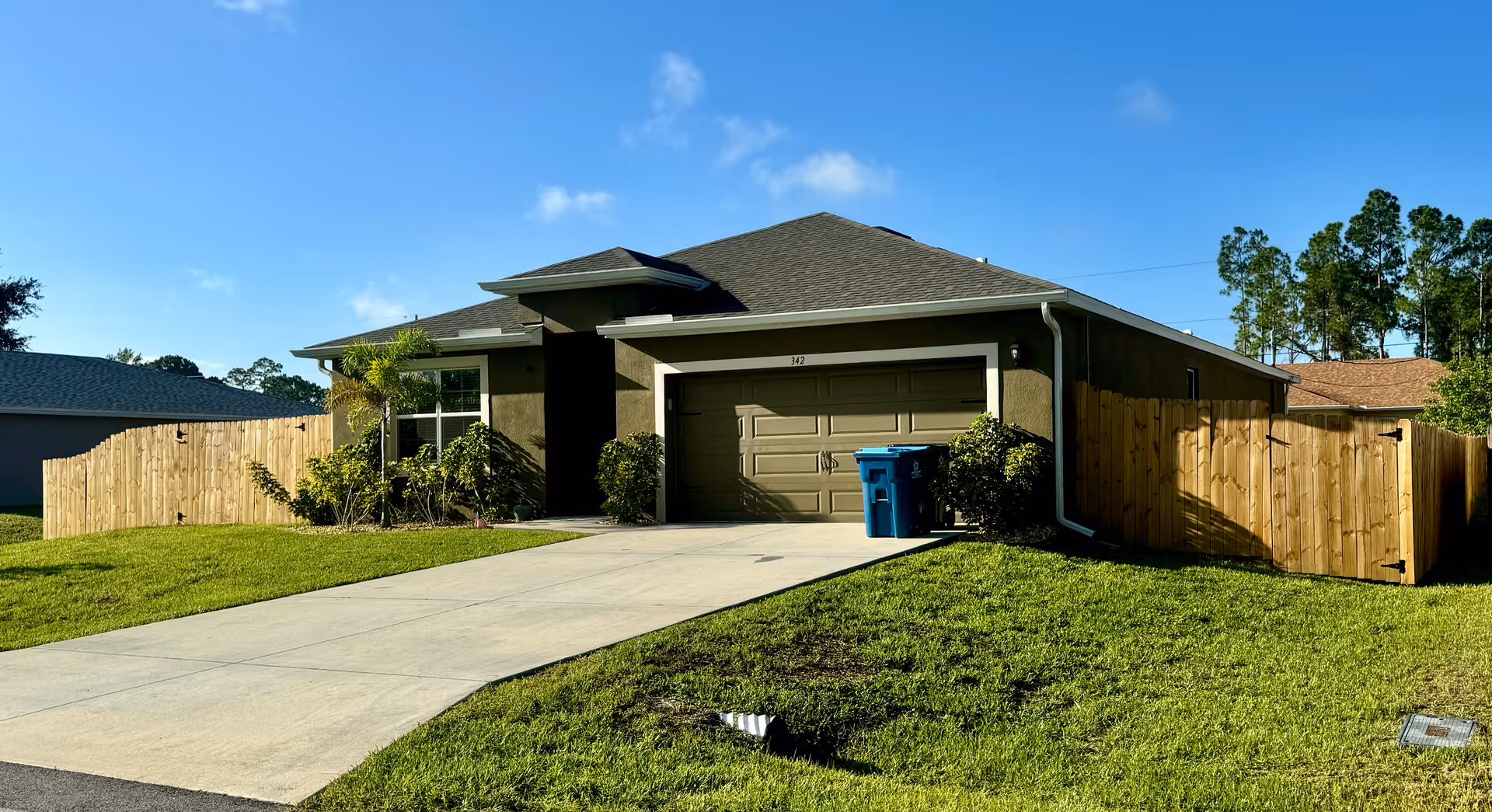 Single-story house with a two-car garage, a concrete driveway, and a well-maintained lawn. The house has a brown exterior with a dark roof and is surrounded by a wooden fence. There are some small bushes and a palm tree near the front window. The sky is clear and blue.