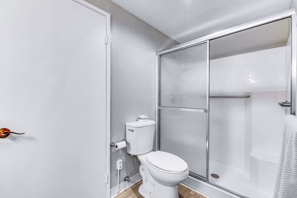 A clean bathroom featuring a white toilet with a roll of toilet paper on the holder and a small decorative item on top of the tank. Next to the toilet is a shower with sliding frosted glass doors and white tiled walls. A towel hangs on a bar attached to the shower door. The walls are painted light gray and the floor has a wood-like finish.