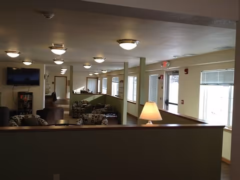 Interior view of a senior living facility common area with multiple seating arrangements including patterned armchairs and sofas. The space is well-lit with ceiling lights and natural light from windows. A table lamp is visible on a partition wall, and a TV is mounted on the far wall above a small bookshelf.