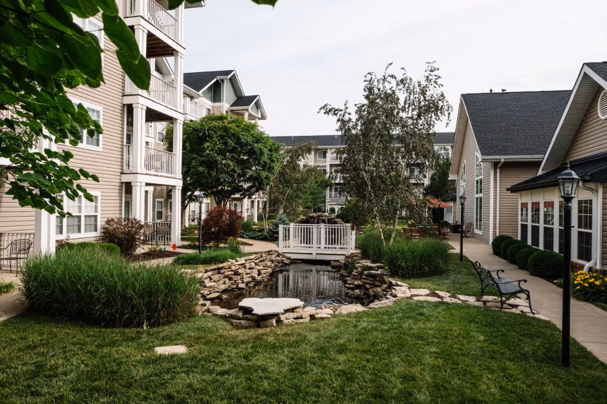Courtyard of a senior living community featuring a small pond with a white footbridge, benches, landscaped lawns and surrounding multi-story buildings.