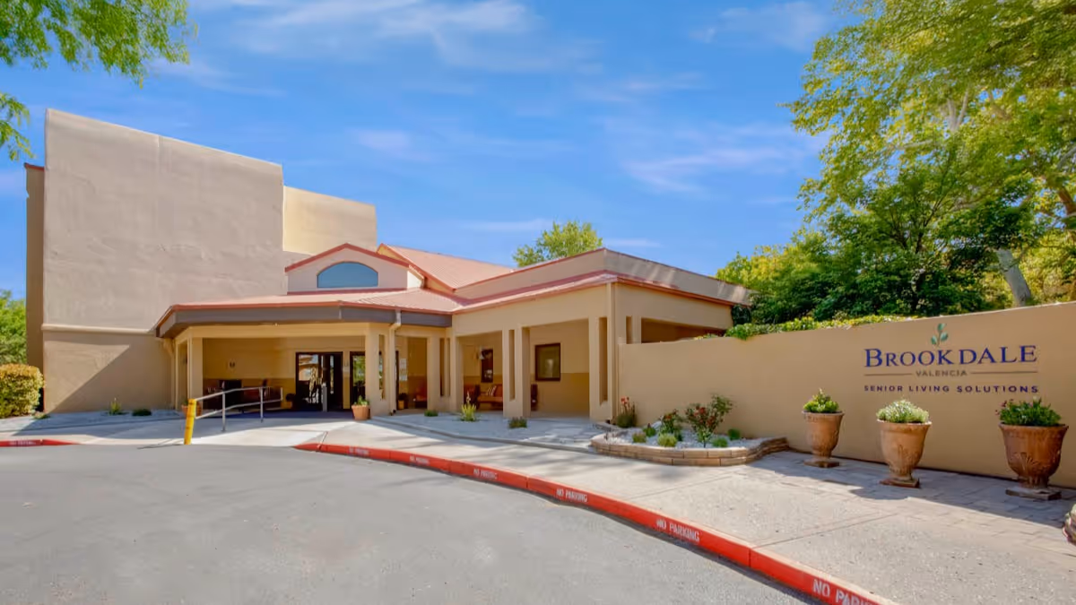 Exterior view of the Brookdale Valencia senior living facility entrance with a covered drop-off area, potted plants, and a sign on the wall reading 'Brookdale Valencia Senior Living Solutions' under a clear blue sky.
