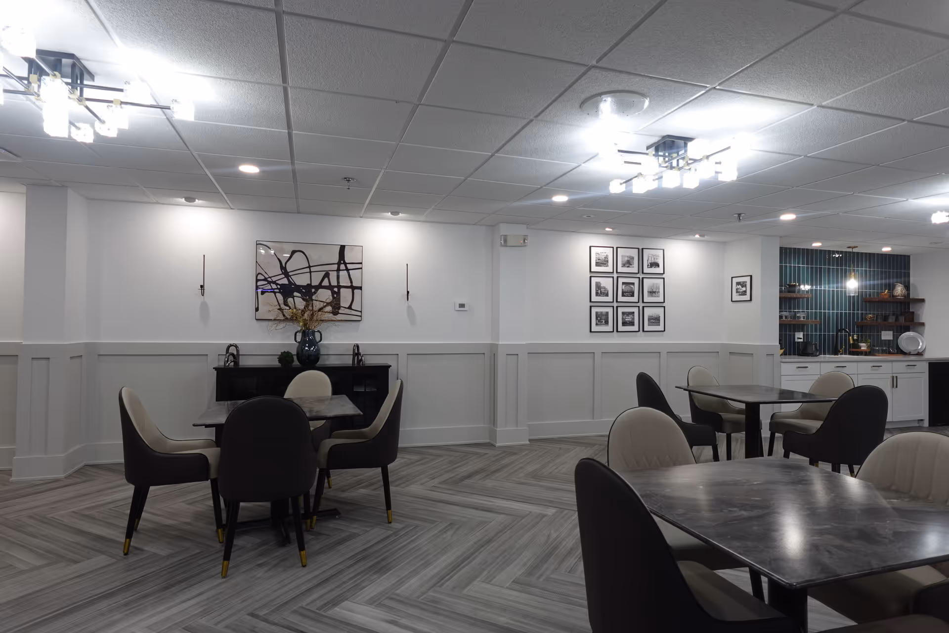 A modern dining area with several tables and chairs arranged neatly. The room features light gray walls with white wainscoting, a patterned gray floor, and contemporary light fixtures on the ceiling. There is abstract artwork and a vase with dried flowers on a black sideboard against the wall, and a small kitchenette area with shelves and a tiled backsplash in the background.