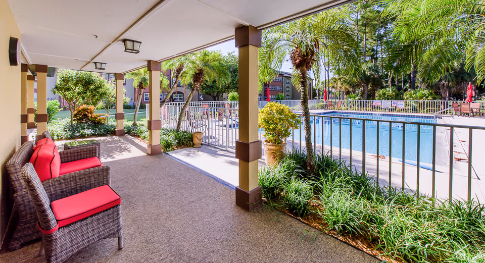 Covered patio area with wicker chairs featuring red cushions overlooking a fenced swimming pool surrounded by palm trees and greenery at Brookdale College Parkway.