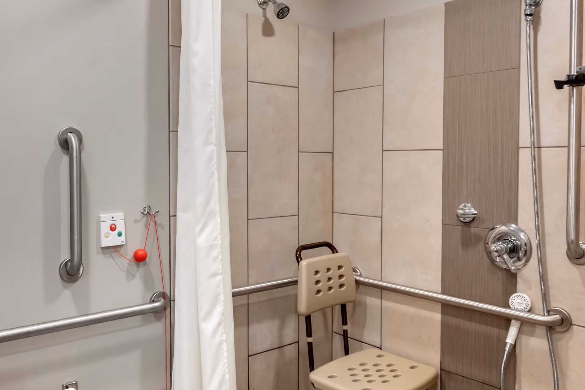 Accessible shower area with beige tiled walls, a beige shower chair with a backrest, stainless steel grab bars on the walls, a handheld showerhead, and an emergency pull cord with a red ball attached.