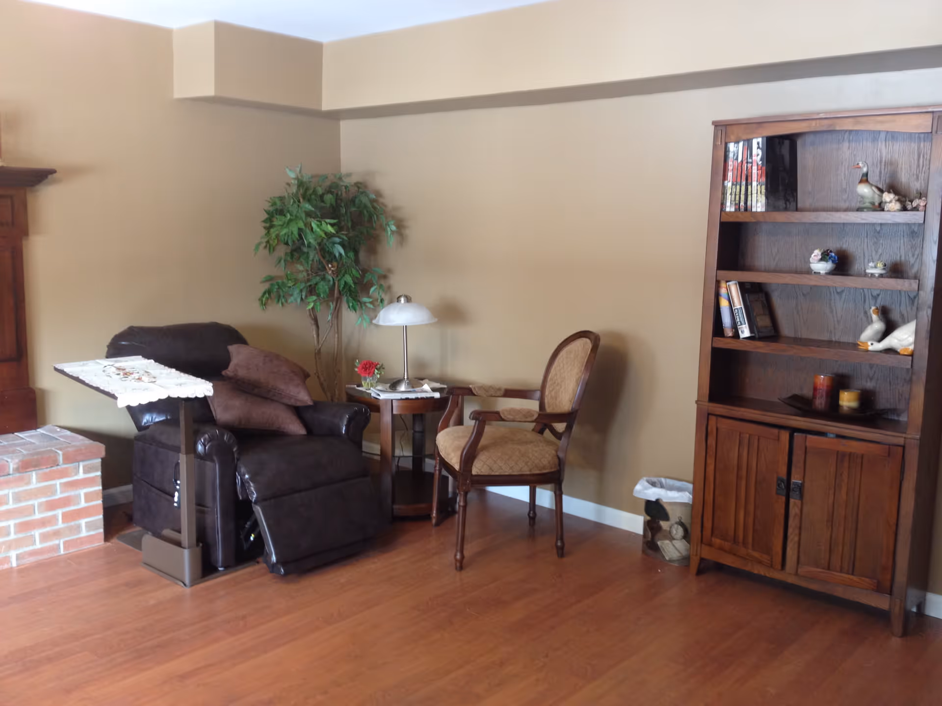 A cozy living room corner with a dark brown recliner chair next to a small wooden side table holding a lamp and a small flower vase. There is a wooden armchair with cushioned seat and backrest beside the table. A tall wooden bookshelf with books and decorative items stands against the wall. The floor is wooden, and there is a brick fireplace partially visible on the left side. A green potted plant is placed behind the recliner chair.