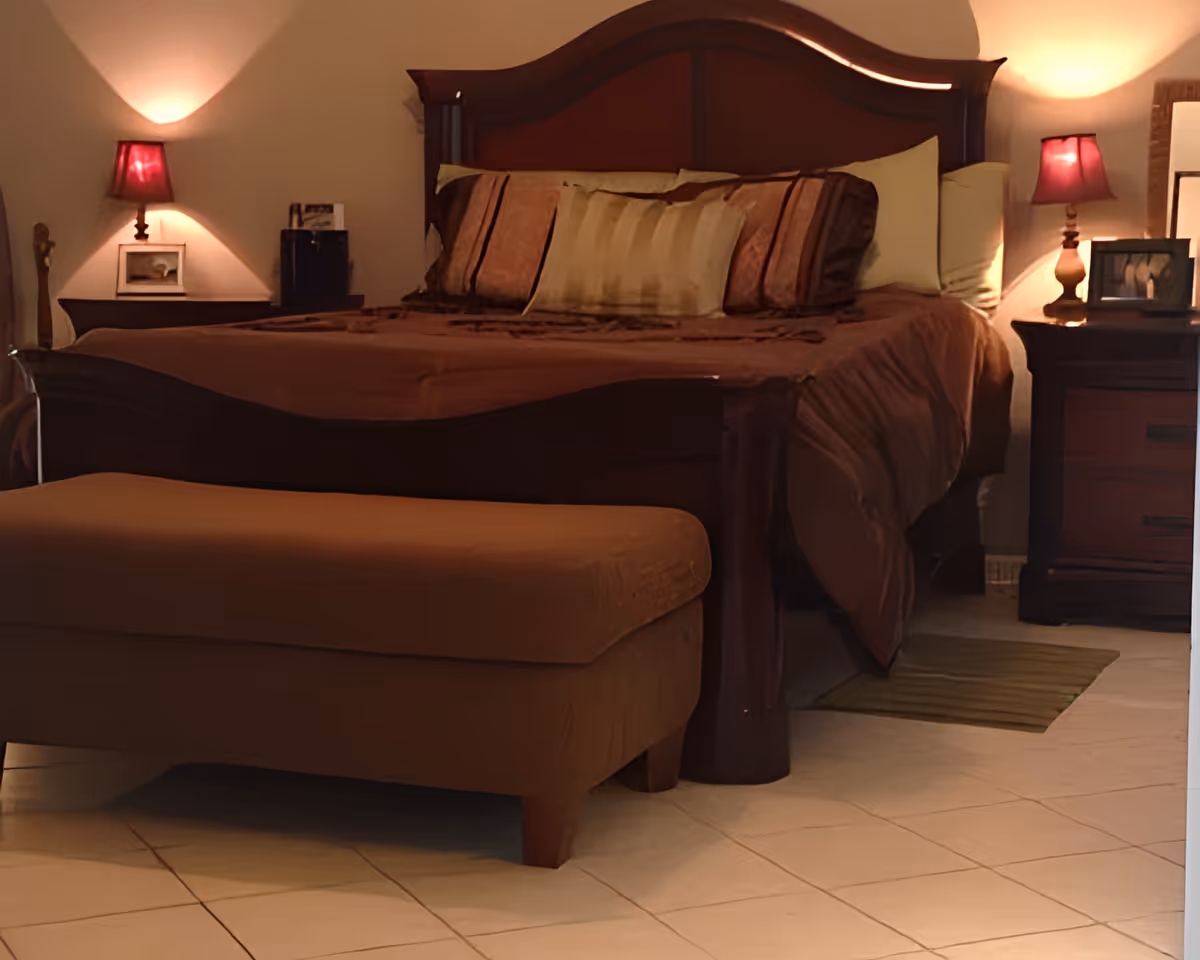 Warmly lit bedroom with a wooden bed, decorative pillows, an upholstered bench at the foot, and bedside tables with lamps.