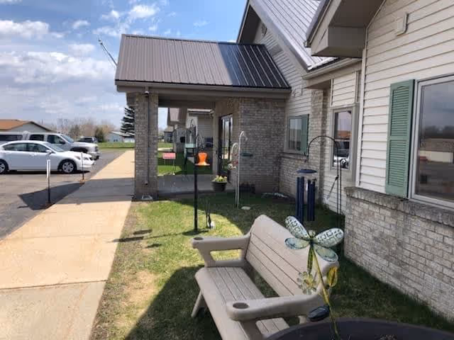 Outdoor view of Vergas Assisted Living facility showing a sidewalk, parked cars, a building with beige siding and green shutters, a covered entrance, a bench with a decorative dragonfly, and some garden decorations on a grassy area.